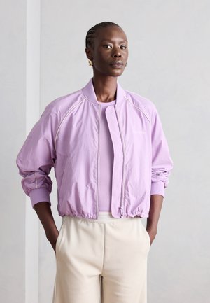 Femme aux cheveux tressés portant des boucles d'oreilles en or, une veste zippée violet clair et un haut, et un pantalon crème taille haute, les mains dans les poches.