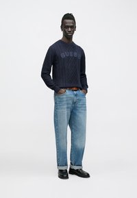 Marineblauer Strickpullover mit Zopfmuster und "GUESS"-Logo, kombiniert mit hellblauen Jeans und schwarzen Schuhen mit Umschlag.