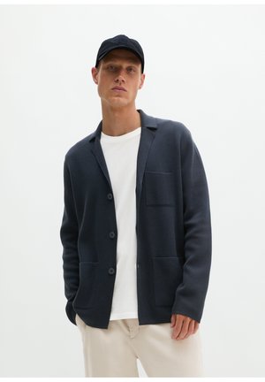 Mann trägt schwarze Baseballkappe, dunkle, geknöpfte Strickjacke mit Taschen, weißes T-Shirt und beige Hose vor schlicht hellem Hintergrund.