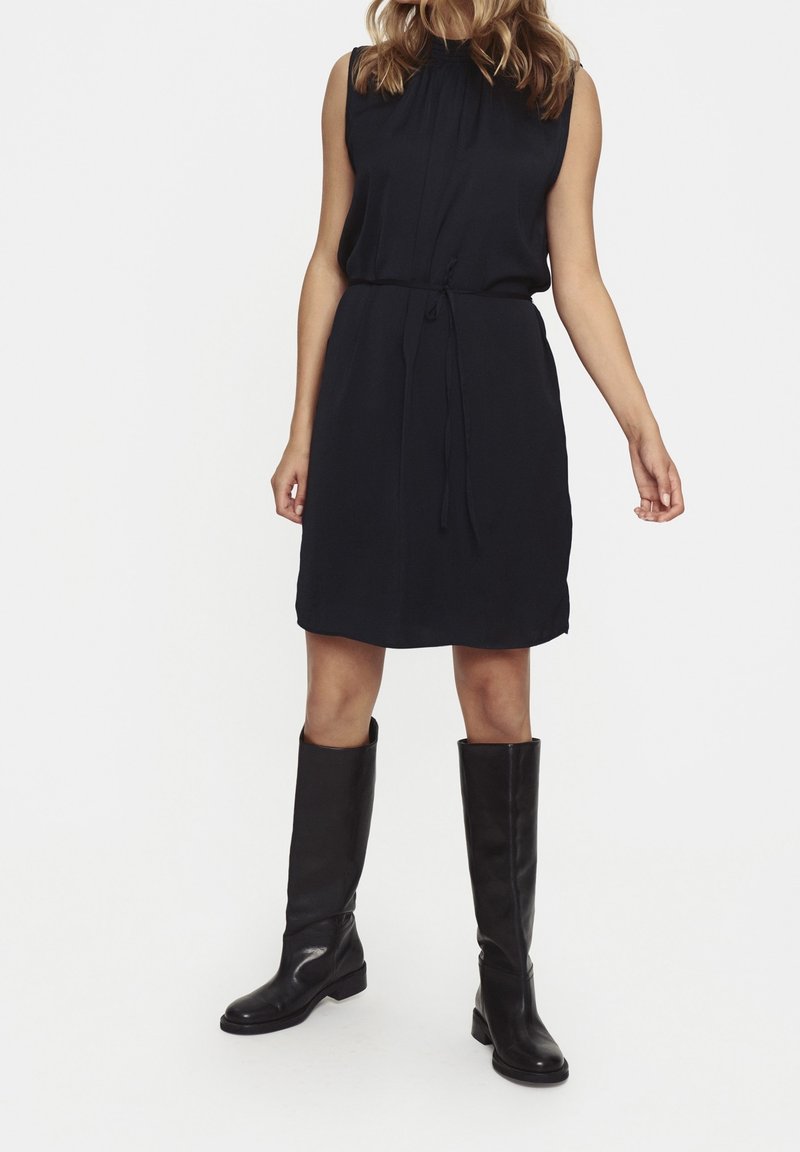 Svart ärmlös klänning med veckad halsringning och dragsnöre i midjan; bärs med knähöga svarta läderstövlar. Slät tyg.