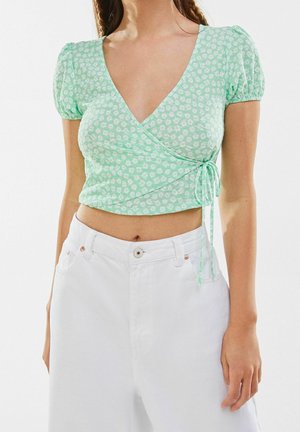 Una donna indossa un top a maniche puff floreale avvolgente in verde chiaro e pantaloni bianchi a vita alta su uno sfondo neutro.