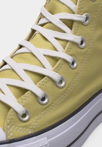 Gelber hoher Canvas-Sneaker mit weißen, kreuzweise geschnürten Schnürsenkeln durch silberne Ösen und einer weißen Gummisohle mit schwarzer Umrandung.