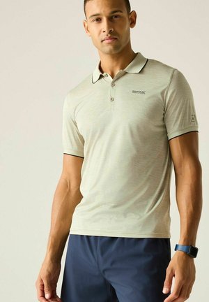 Hombre con camiseta polo gris clara y pantalones cortos azul marino, de pie con expresión neutra contra un fondo sencillo.