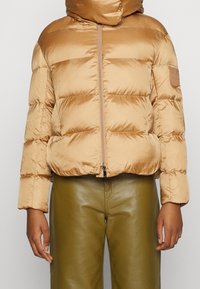 Giacca puffer color sabbia con colletto alto e chiusura a zip, caratterizzata da cuciture orizzontali, texture liscia e una patch con logo sulla manica.