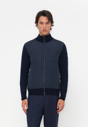 Navy zip-up jacket with a quilted front and knitted sleeves. Features a high collar and ribbed waistband. Simple design without patterns.