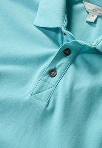 Ljusblå polo skjorta av texturerad ekologisk bomull, med en treknapsknäppning med bruna knappar och en vikt krage.