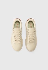Beige sneakers met een gladde textuur, afgeronde neuzen, contrasterende rode accenten op de hiel en beige veters over een minimalistisch ontwerp.