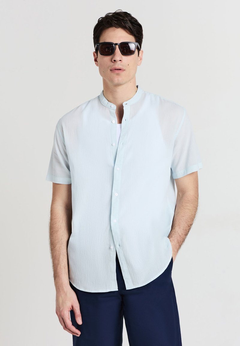 Uomo che indossa una camicia a maniche corte azzurro chiaro con bottoni, occhiali da sole neri e pantaloni blu scuro, in piedi contro uno sfondo bianco semplice.