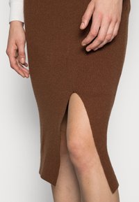 Hnědá pletená sukně s žebrováním a bočním rozparkem, texturovaná tkanina, délka ke kolenům a přiléhavý střih. Ruka položená na sukni.