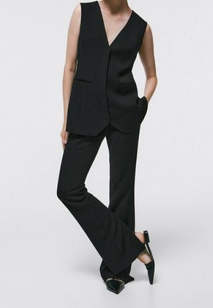 Mannequin portant un gilet noir sans manches sur mesure avec un pantalon évasé noir assorti et des chaussures slingback noires à bouts pointus, debout les mains dans les poches.