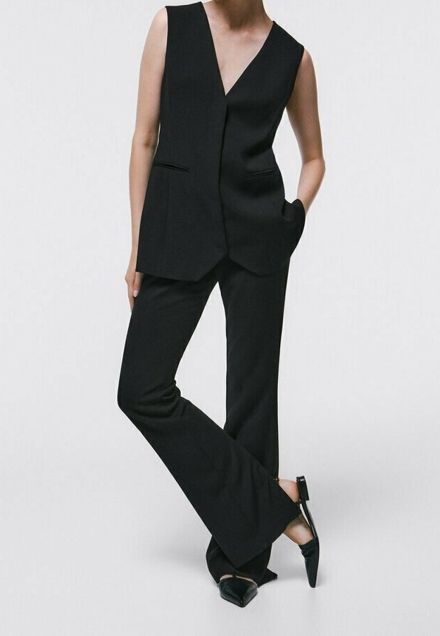 Mannequin portant un gilet noir sans manches sur mesure avec un pantalon évasé noir assorti et des chaussures slingback noires à bouts pointus, debout les mains dans les poches.