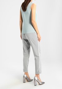 Kvinna iklädd en ljusblå ärmlös topp, grå byxor med vita sidostripes och metalliska silver blockklackskor, stående med ryggen vänd framåt.