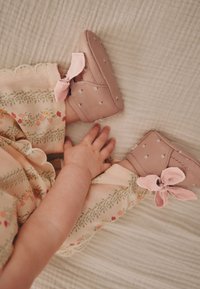 Bébé portant des chaussures roses souples avec de petits motifs floraux et des nœuds, vêtu d’un pantalon à motifs floraux sur une surface claire texturée.