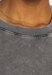 Primo piano del collo di una persona che indossa una sottile collana di oro e un maglione grigio a coste con collo rotondo.