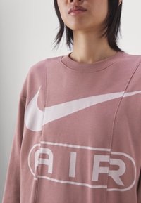 Růžová mikina vyrobená z bavlny, s velkým bílým logem Nike a grafickým potiskem "AIR". Hřejivý kulatý výstřih a volný střih.
