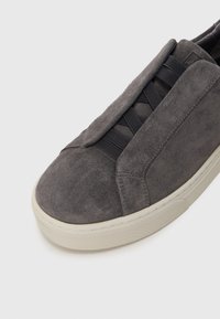 Zapatilla de deporte tipo slip-on de ante gris con cordones elásticos, punta redondeada y suela de goma blanca texturizada. Superficie suave y diseño minimalista.