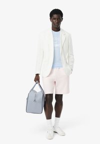 Blazer de lino blanco sobre una camiseta con logo azul claro, pantalones cortos rosas y bolso gris. El modelo lleva zapatillas blancas y calcetines blancos.