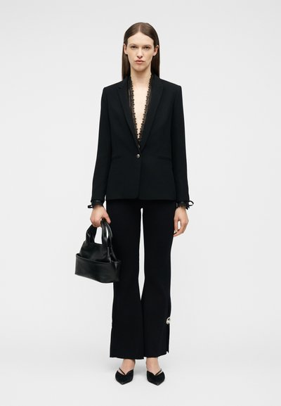 Γυναίκα που φοράει μαύρο ραμμένο σακάκι με δαντελένιο περίγραμμα, μαύρο καμπάνα παντελόνι με ασημένια κουμπιά, μαύρες μυτερές γόβες, κρατώντας μαύρη τσάντα.