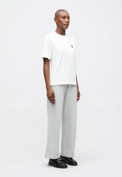 Λευκό βαμβακερό T-shirt με στρογγυλή λαιμόκοψη και κοντά μανίκια, συνδυασμένο με ανοιχτό γκρι βαμβακερό παντελόνι φόρμας. Μαύρα δερμάτινα παπούτσια με κορδόνια ολοκληρώνουν την εμφάνιση.