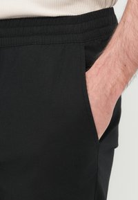 Μαύρο παντελόνι με ελαστική μέση, πλαϊνές τσέπες και λεία υφή. Κοντινή λήψη που αναδεικνύει τις λεπτομέρειες του υφάσματος και τη θέση των τσεπών.