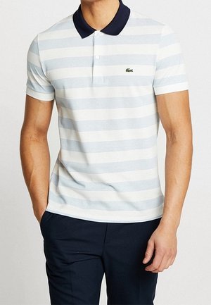 Homme portant un polo rayé blanc et bleu clair avec un col marine et un pantalon bleu marine, debout les mains dans les poches.