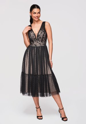 Kobieta w czarnej koronkowej sukience bez rękawów z głębokim dekoltem w kształcie litery V i prześwitującą spódnicą w kropki, stojąca z jedną ręką przy ramieniu.