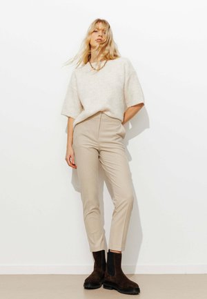 Pull en tricot beige clair, pantalon beige taille haute et bottines marron avec accents noirs. Texture lisse et coupe ajustée.