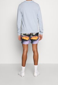 Světle modrý dlouhý svetr s žebrovanými manžetami doplněný barevnými kraťasy s abstraktním vzorem v purpurové, oranžové a černé barvě. Viditelné bílé tenisky.