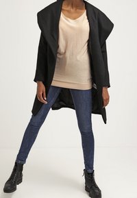 Lungo cappotto nero con ampi rever, indossato sopra un top beige chiaro. La modella indossa jeans blu scuri e stivali neri con lacci.