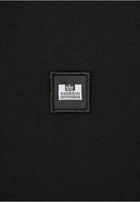 Sort tekstil med et kvadratisk patch med "WEEKEND OFFENDER" i hvid tekst og et krone-logo, syet med en sort kant.