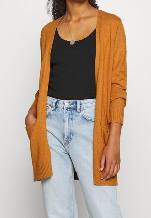 Bruine gebreide cardigan met een open voorkant, geribbelde boorden en twee voorzakken, gecombineerd met een zwarte tanktop en lichtblauwe jeans.