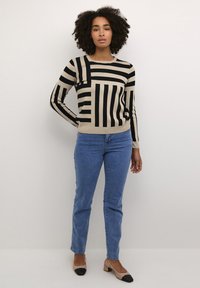 Pull en beige et noir à rayures avec des manches longues, associé à un jean bleu et des chaussures à talons bloc noir et beige. Texture lisse, design ajusté.