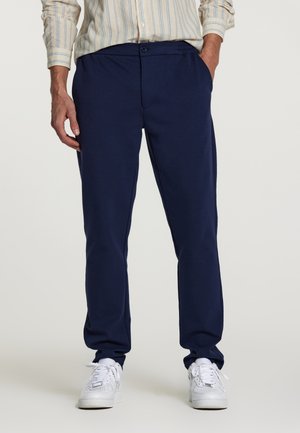 Pantaloni de sport de culoare albastru închis, cu o croială lejeră, dotati cu buzunare înclinate și închidere cu nasture la talie. Purtați cu adidași albi.