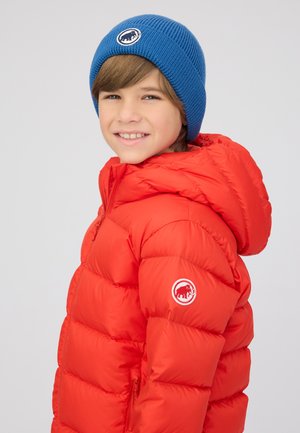 Rote Steppjacke mit Kapuze, ausgestattet mit einem diagonalen Steppmuster und einem gestickten Logo auf der Schulter. Blau gestrickte Mütze rundet den Look ab.