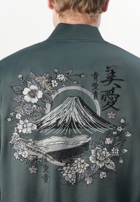 Green bomber jacket with detailed floral and mountain embroidery on the back, featuring black and silver threads, and a smooth texture.
