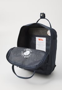 Mörkblå ryggsäck med lock, med en logopatch, tygtextur och en grå foder. Inuti synlig etikett med skötselråd.