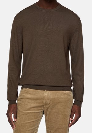 Mand iført en langærmet brun crewneck sweater og lysebrune ribbukser, stående med armene afslappet ned langs siden.