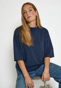 Haut surdimensionné bleu marine avec un col montant et des manches larges, associé à un jean bleu clair. Le mannequin est assis sur un tabouret devant un fond uni.