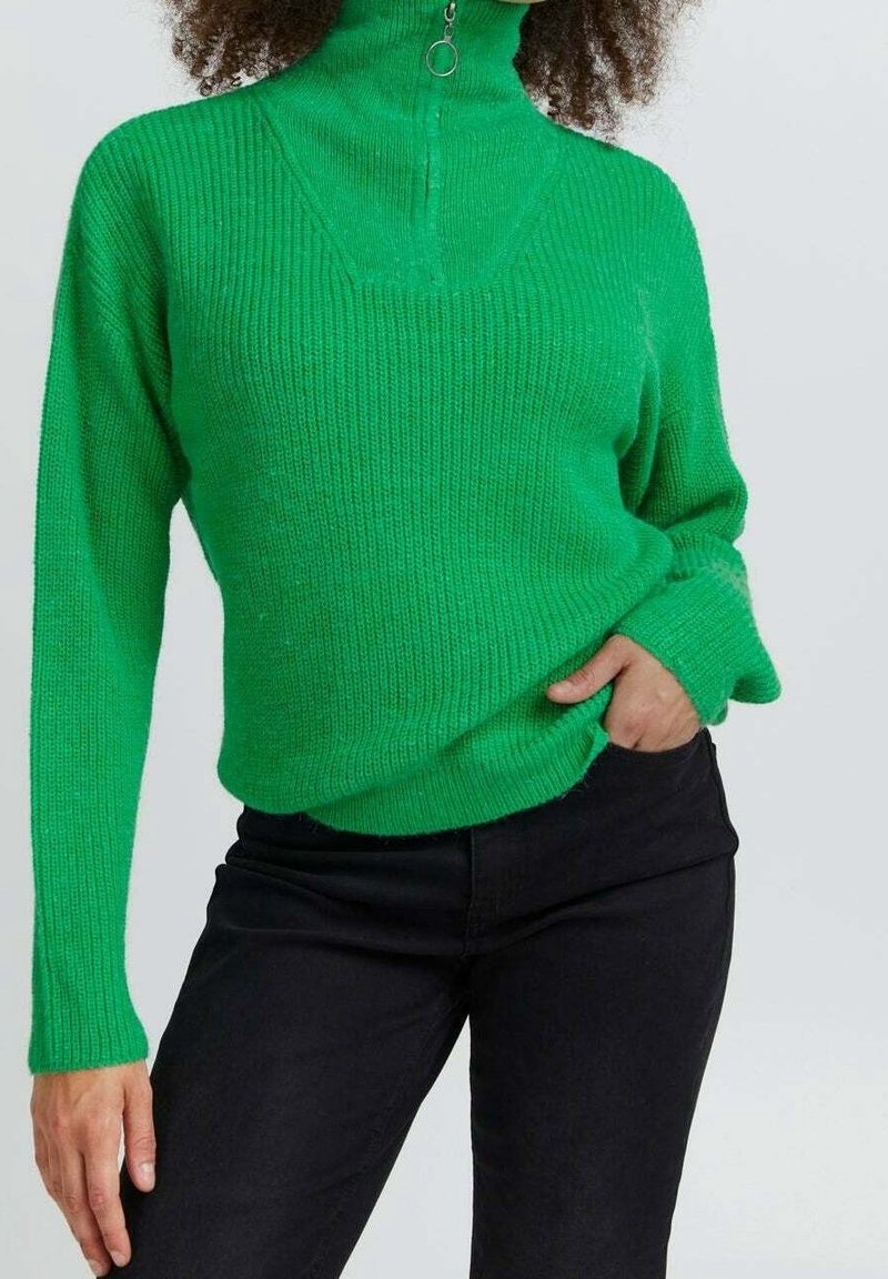 Grüner, gerippter Strickpullover mit hohem Kragen und Reißverschluss. Verfügt über lange Ärmel und eine lockere Passform. Kombiniert mit schwarzen Hosen.