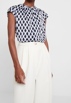 Vrouw draagt een mouwloze zwart-wit geometrisch patroon blouse, gestopt in een witte, hooggetailleerde broek met wijde pijpen.
