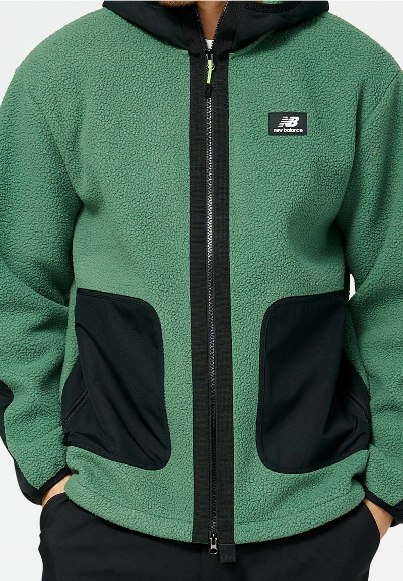 Groene fleece jas met grote zwarte voorzakken, zwarte ritsvoering, New Balance-logo op de borst, gedragen met handen in de zakken.