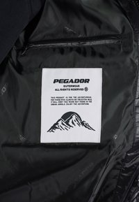 Bílá nášivka značky PEGADOR přišita uvnitř podšívky černé bundy s grafickým zobrazením hory a textem o dobrodružství a teple.