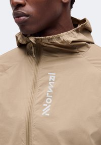 Gros plan d'une personne portant une veste à capuche beige avec une fermeture éclair à l'avant et un logo texte blanc vertical sur la poitrine.