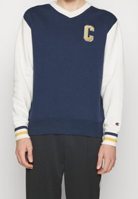 Námořnicky modrá a bílá mikina s výstřihem do V, zdobená vyšívaným zlatým logem "C" a pruhem na manžetách v námořnicky modré, bílé a žluté barvě.