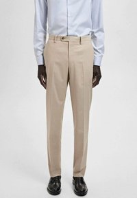 Pantaloni beige su misura con taglio dritto, due pieghe frontali e passanti per cintura, abbinati a scarpe in pelle nere. Tessuto dalla texture liscia.