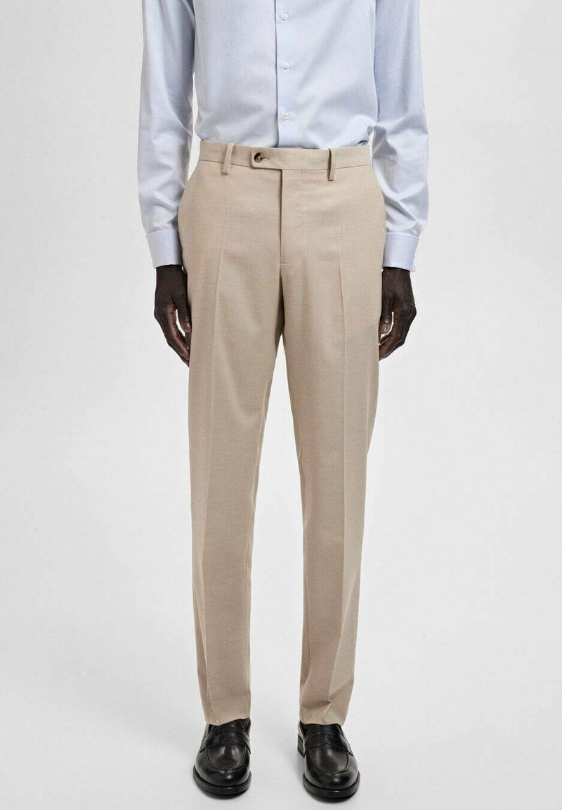 Pantaloni beige su misura con taglio dritto, due pieghe frontali e passanti per cintura, abbinati a scarpe in pelle nere. Tessuto dalla texture liscia.