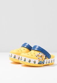 Gula träskor med blå rem, med ett mönster av små hål och grafik av Minions runt den vita basen. Mjuk plastmaterial.