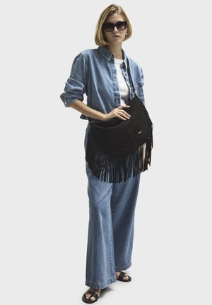 Black suede handbag with fringe detailing. Model wears a light denim shirt, white t-shirt, and wide-leg denim pants. Dark sunglasses complete the look.