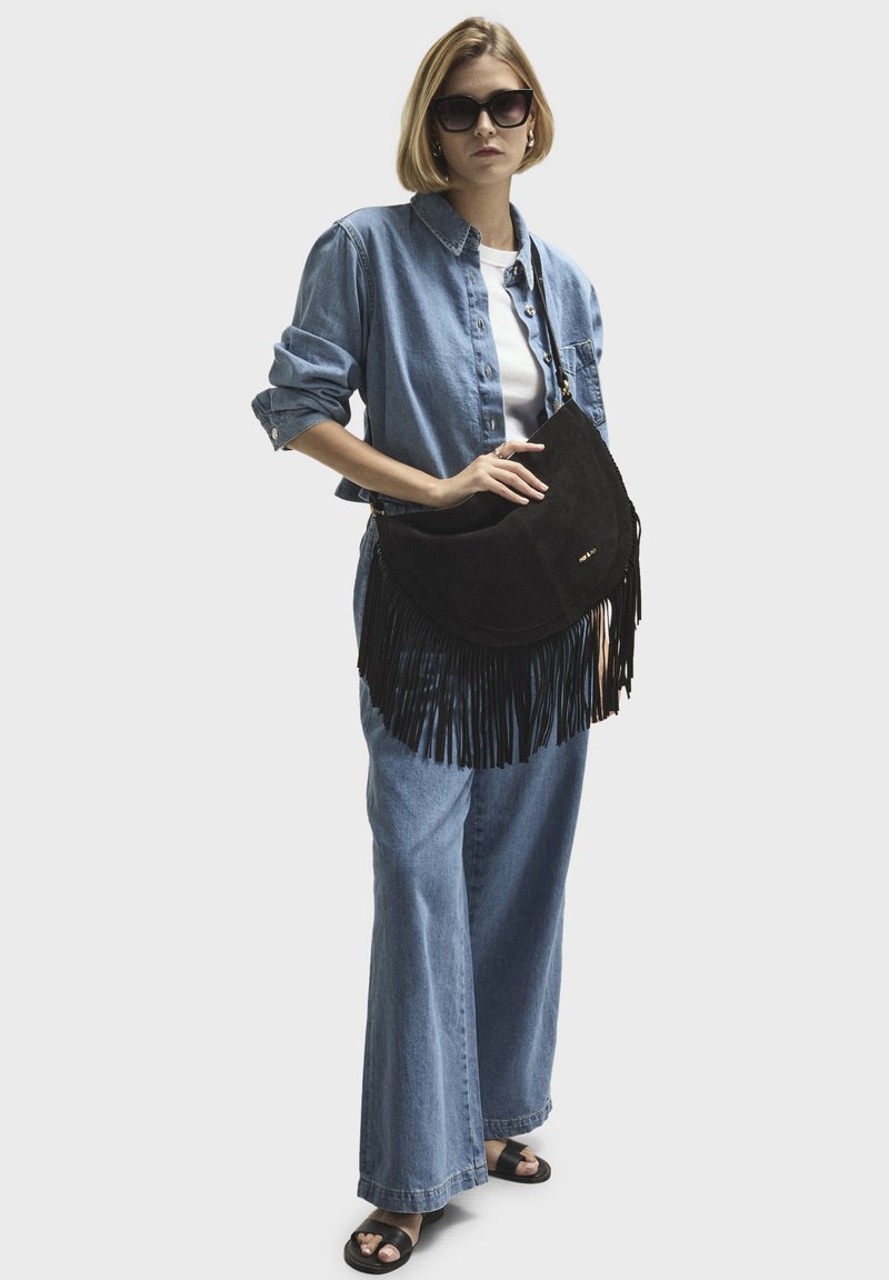 Black suede handbag with fringe detailing. Model wears a light denim shirt, white t-shirt, and wide-leg denim pants. Dark sunglasses complete the look.