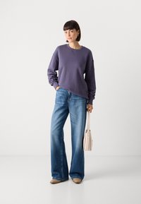 Violetinis džemperis su apvaliu iškirpimu, derinamas su plačiomis mėlynomis džinsinėmis kelnėmis. Modelis laiko mažą šviesiai beigę rankinę. Lygus audinio paviršius.
